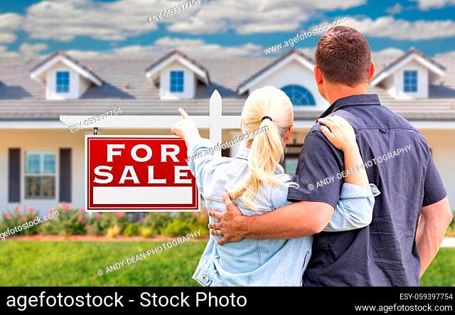 Young Adult Couple Facing and Pointing to Front of For Sale Real Estate Sign and House