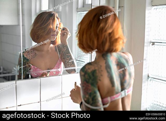 Concentrated girl with colorful tattoos applying mascara on her eyelashes while standing opposite the mirror in the bathroom with white tiled walls