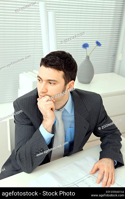Architect wearing grey suit sitting at office desk, thinking over floor plan. Overhead shot
