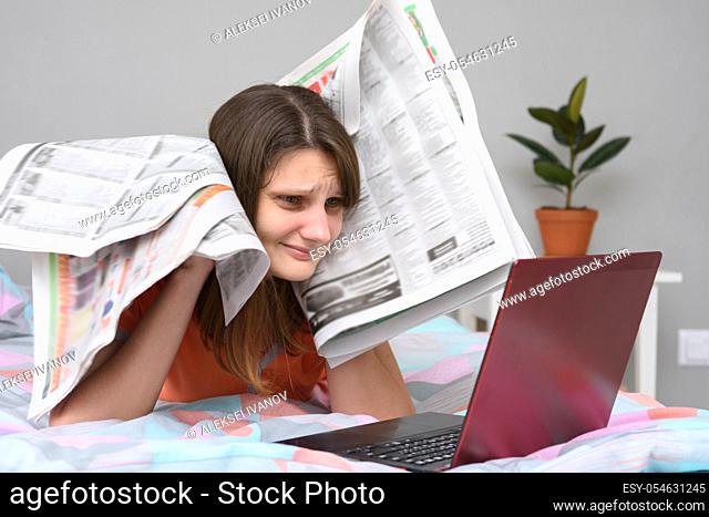Girl put newspapers to her head and looks at the laptop screen trying to find work