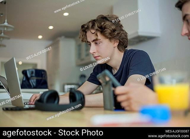 Two teenage boys using smartphone and laptop on table at home