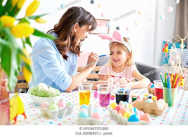 Woman painting daughter's nose while painting easter eggs at table
