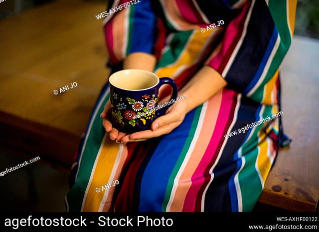 Woman in multi colored clothing holding coffee cup while sitting on table at home