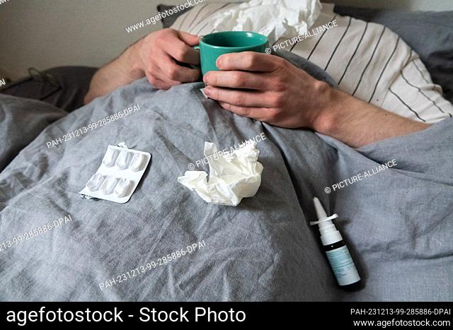 12 December 2023, Lower Saxony, Hanover: ILLUSTRATION - A man lies in bed with handkerchiefs, teacup, nasal spray and tablets (staged scene)