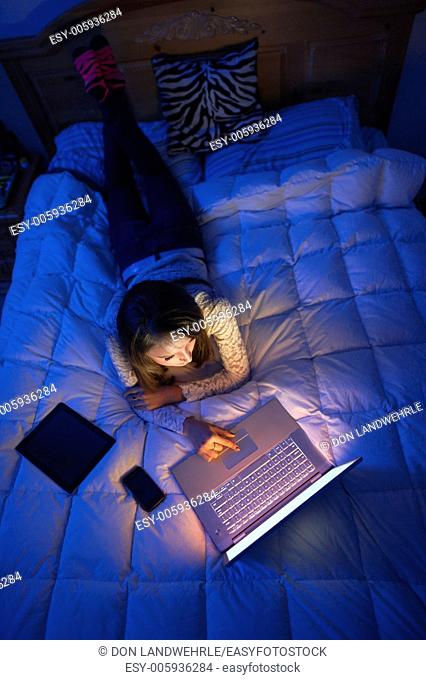 Teenage girl lying on a bed at night working on a laptop computer