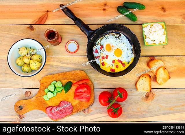Different food: scrambled eggs in frying pan, boiled potatoes, curd, croutons, radishes, cucumbers, tomatoes, smoked sausage, mint, sorrel on wooden table