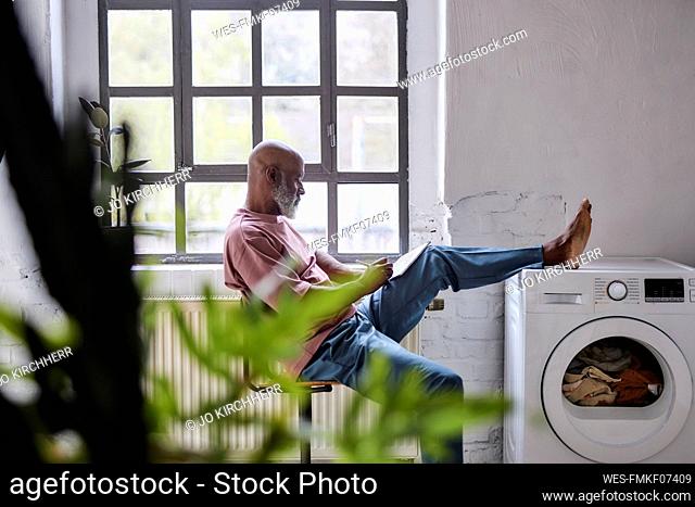 Bald man using tablet PC keeping foot on washing machine at home