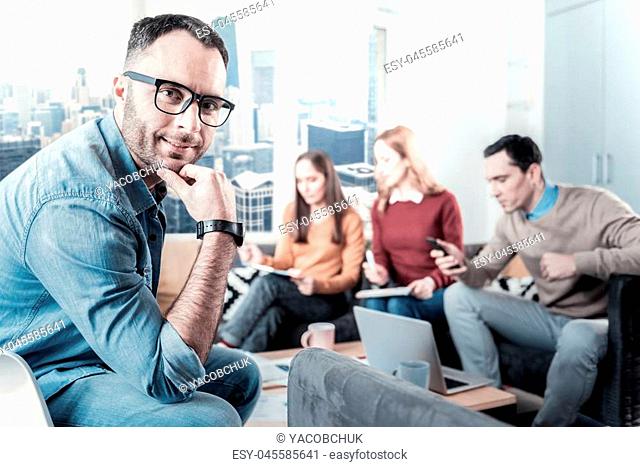 Always ready. Pleasant bespectacled handsome man sitting on the chair near the table holding his right hand near head and looking straight