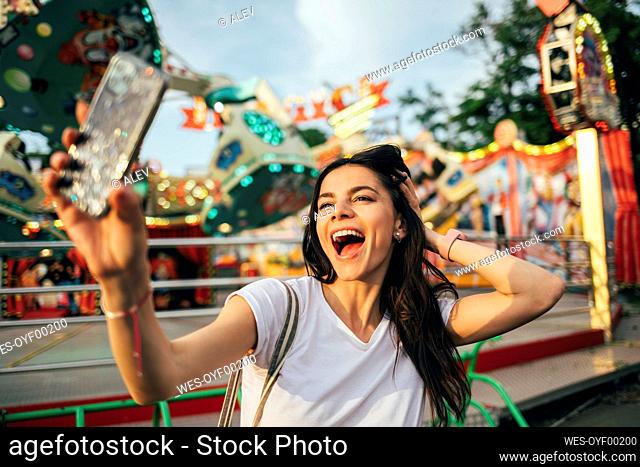 Cheerful young woman with hand in hair taking selfie at amusement park