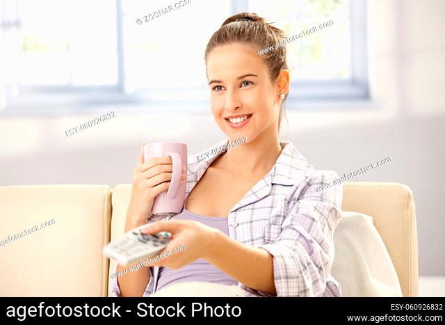 Laughing woman watching tv, using remote control, having morning tea on couch in pyjama