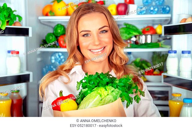 Vegetarian lifestyle, portrait of a beautiful woman standing near open fridge full of different fresh vegetables, organic nutrition, weight control
