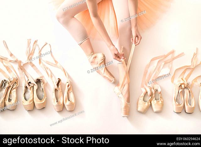 Ballet dancer sits on the white floor and dresses a beige pointe shoe in the studio. She wears a light dance wear and a peach tutu