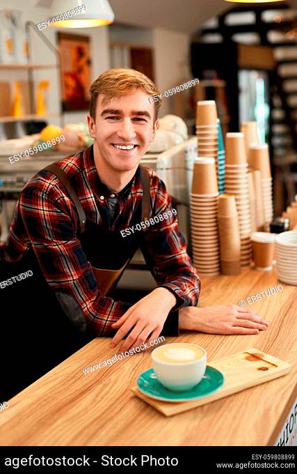 smiling male waiter in apron with cup of hot drink in coffee shop, barista is happy to work in the cafeteria