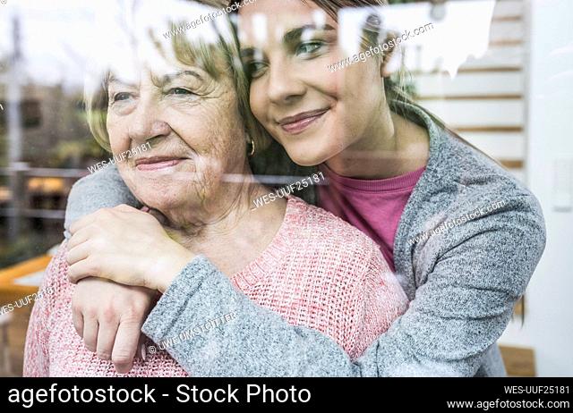 Young caregiver hugging senior woman looking out from window