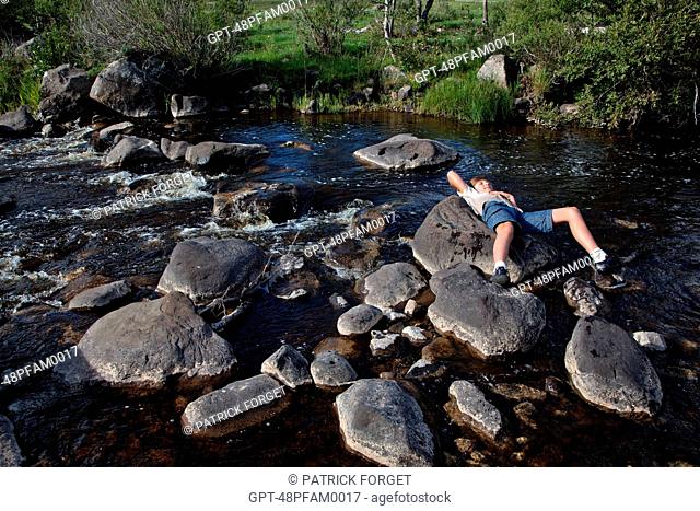 CHILD LYING ON THE STONES IN THE MIDDLE OF A RIVER, NASBINALS, LOZERE 48, AUBRAC, FRANCE