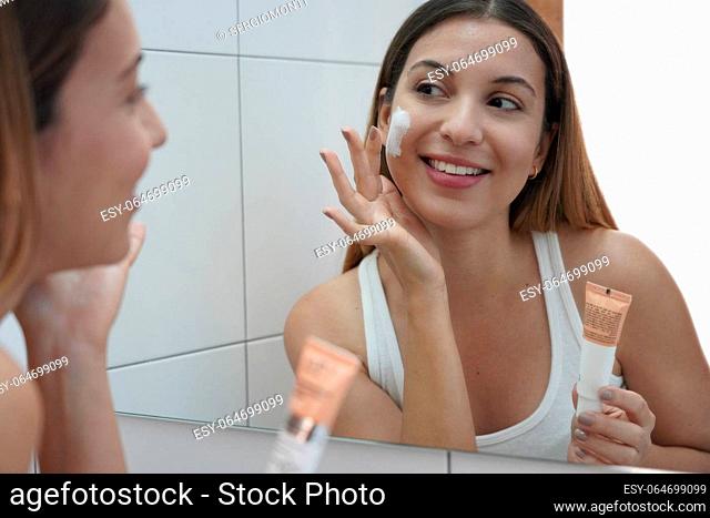 Daily routine. Smiling young woman looking at mirror using face cream