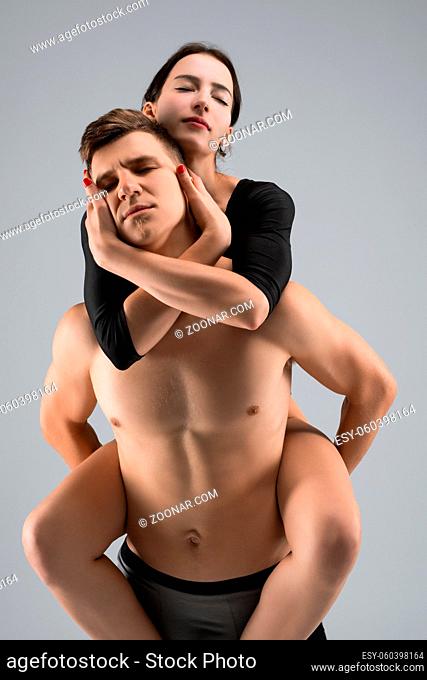 Strong shirtless man with closed eyes carrying barefoot woman on back against gray background