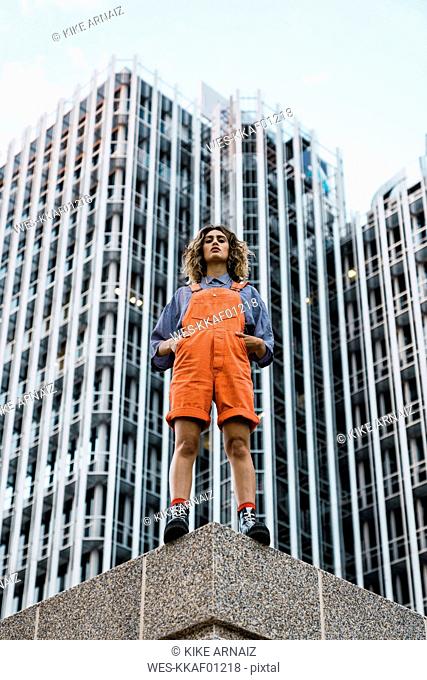 Beautiful woman wearing dungarees, standing on ledge in front of modern high-rise building