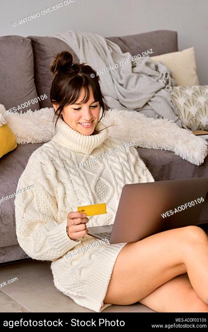 Smiling woman shopping online with credit card through laptop while sitting in living room at home
