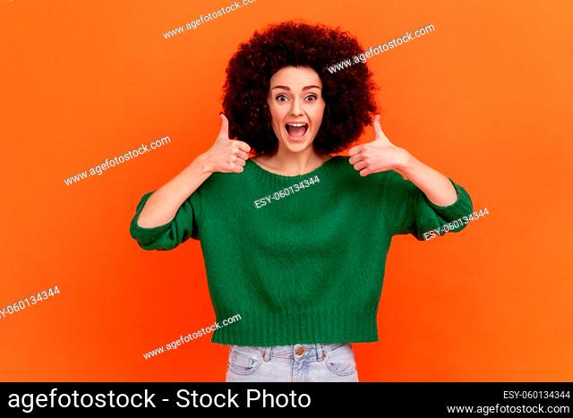 Amazed positive woman with Afro hairstyle wearing green casual style sweater standing showing thumb up with both hands, recommend content