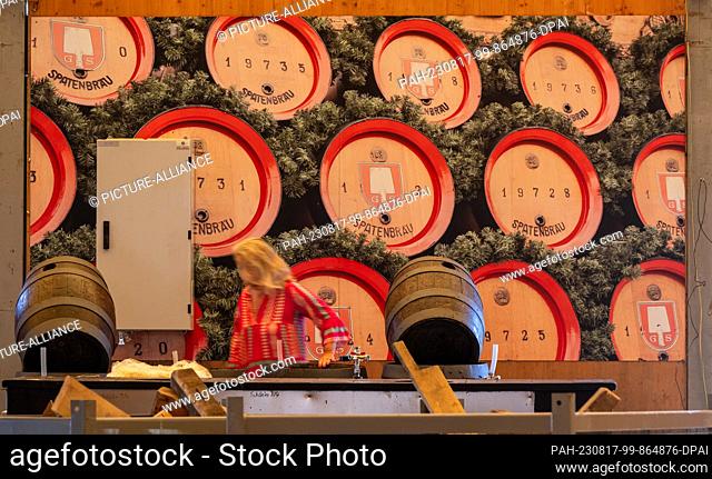 17 August 2023, Bavaria, Munich: A woman walks past a picture of beer barrels on the Oktoberfest grounds that hangs in a beer tent at the still unfinished bar...