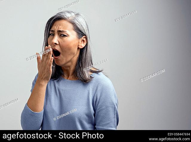 Attractive Asian Mature Woman Yaws and Covers Mouth with Hand in Tiredeness or Boredom. Hair are Behind Ear. She Wears Light-Blue Sweater