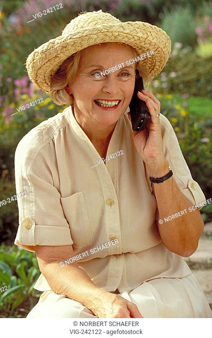 portrait, half-figure, woman mid of 60 wearing beige blouse, skirt and a strawhat sits in the garden making a call with her mobile  - GERMANY, 23/08/2004