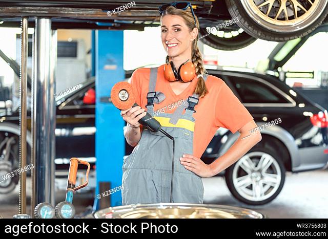 Portrait of a cheerful female auto mechanic wearing safety equipment while holding a car polishing machine in a modern automobile repair shop