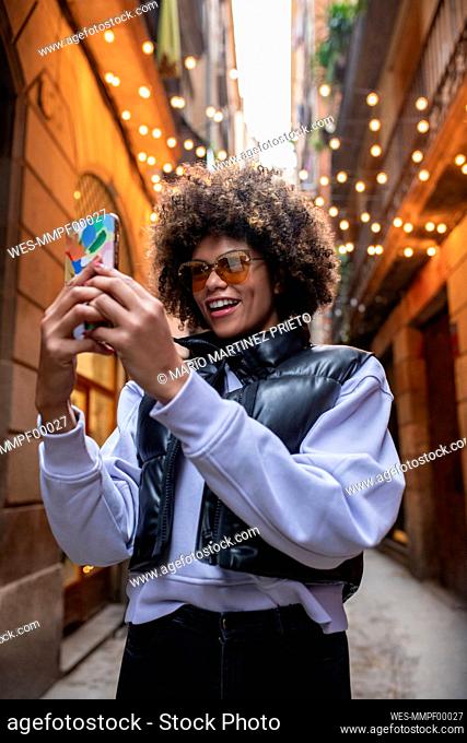 Happy young woman wearing sunglasses taking selfie through smart phone in alley