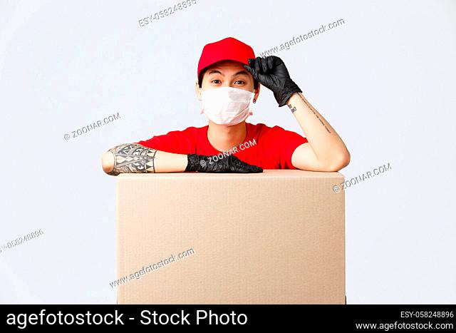 Friendly-looking asian delivery guy in red cap and t-shirt, courier service lean on cardboard box or client package, saluring as making order