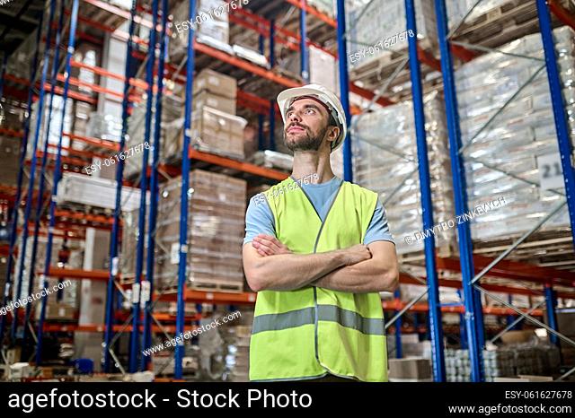 Calm focused storehouse worker in a helmet standing among the shelves with cargo boxes and looking up