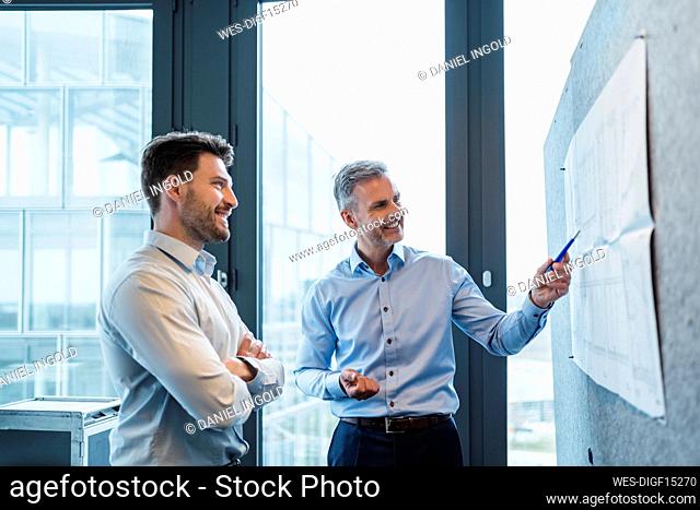 Male professionals discussing over paper on bulletin board at work place