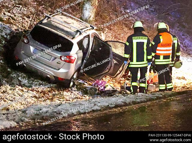 29 November 2023, Saarland, St. Wendel: Firefighters stand in front of a wreck in front of a tree. A 27-year-old driver has died after crashing into a tree near...