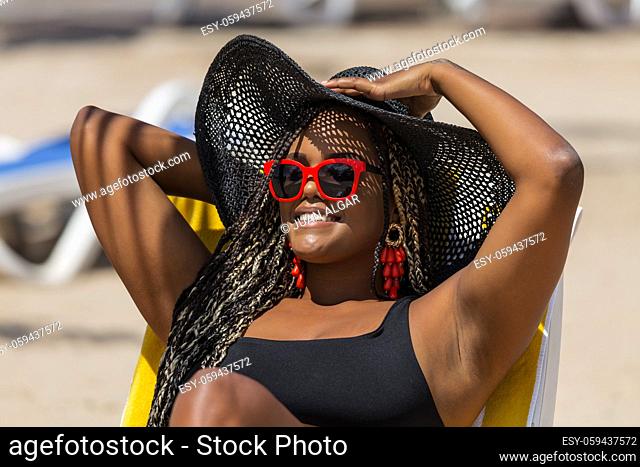 Close-up of an African-American woman on the beach lying on a deck chair wearing a black hat and sunglasses. Cheerful adult woman enjoying a summer day at the...