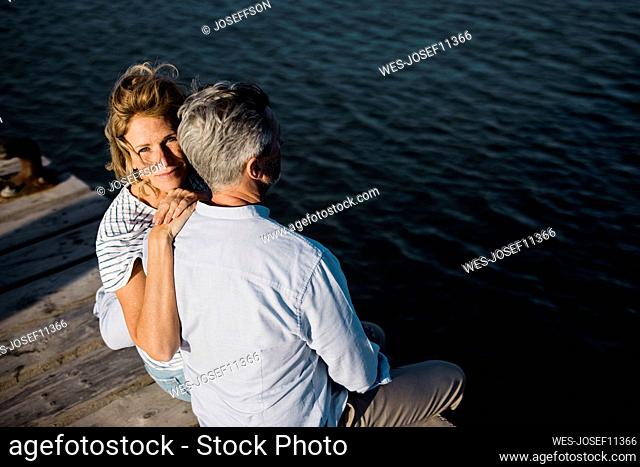 Smiling mature woman sitting with hand on man's shoulder at jetty