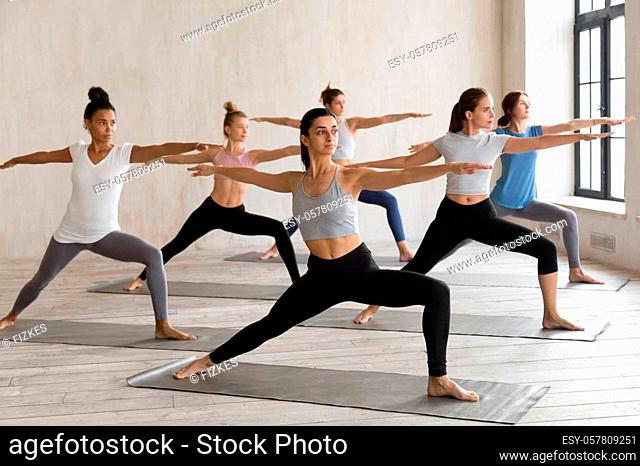 Indian girl and diverse group of young sporty people practicing yoga, doing Warrior II exercise, Virabhadrasana 2 pose, working out, indoor full length
