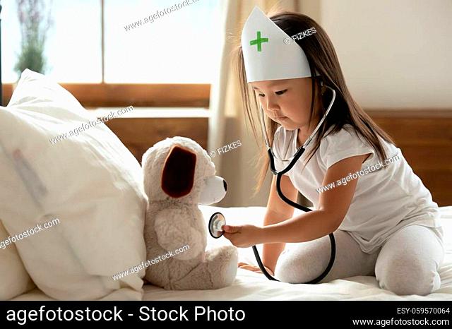 Asian little girl wear medical cap uniform hold phonendoscope listens heartbeat to stuffed toy dog heals best friend takes care of him, healthcare, pediatrician