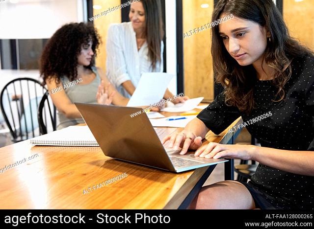 Young woman working on laptop with businesswomen discussing in background