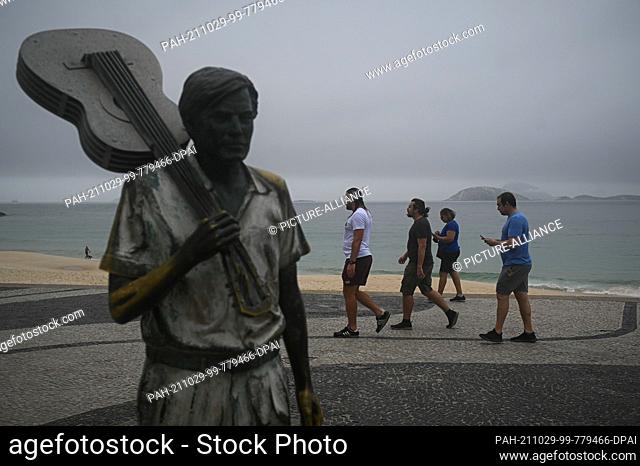 28 October 2021, Brazil, Rio De Janeiro: People without mouth-nose protection walk next to the statue of composer Tom Jobim on Ipanema beach