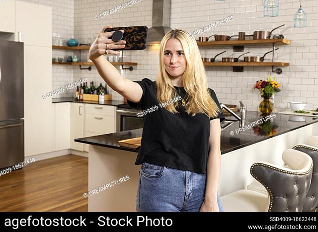 Young adult woman taking selfie while standing in her new apartment with kitchen in background