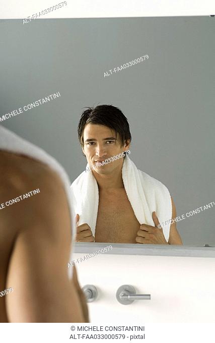 Bare-chested man looking in mirror, smiling at camera, towel slung across shoulders