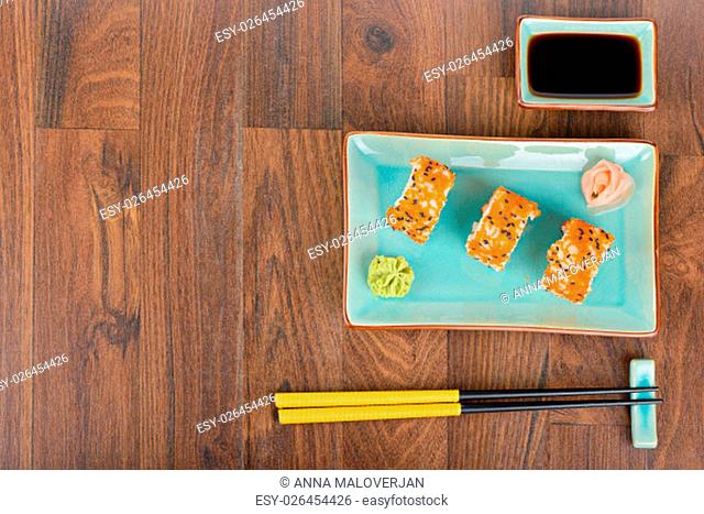 Sushi rolls with masago, served on turquoise plate with pickled ginger, soy sauce and chopsticks on wooden table. Overhead view