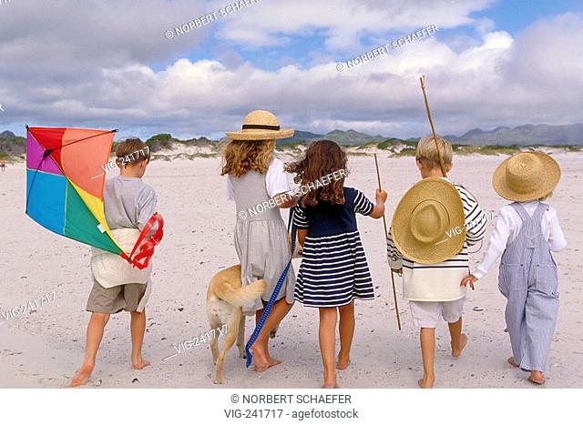 beach-scene, group of 5 children in the age of 7-10 years viewed from the back walking with a dog and a coulered kite, strawhats and sticks bare feeted along...