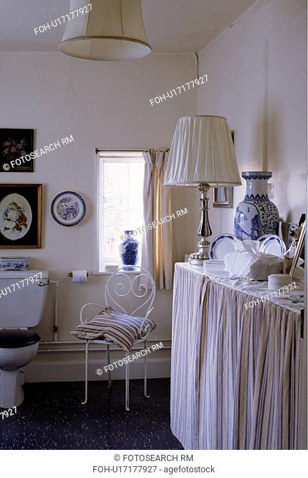 Striped cushion on metal chair in white cottage bathroom with lamp on vanity unit with blue+white striped curtains