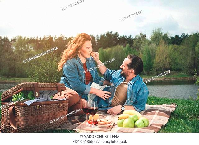 Cheerful man is feeding his wife by small nut and smiling. Couple is having picnic in the nature