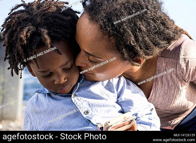 loving african american mother kissing her little son watching video or playing game using mobile phone outdoors. caring mother kissing her little son outdoors...