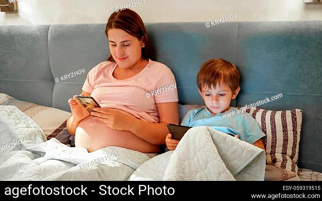 Young pregnant mother with little son lying in bed and using smartphone and tablet computer before going to sleep at night