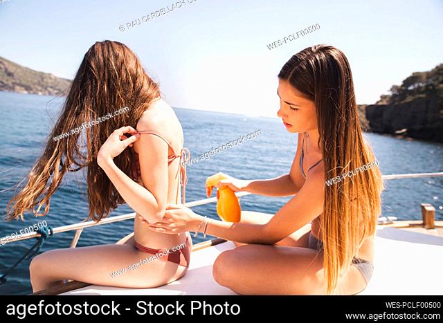 Woman spraying suntan lotion on teenage friend sitting on boat deck