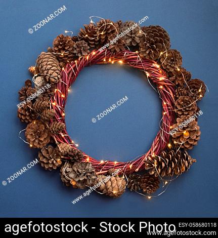 Christmas wreath of pine cones and glowing lights garland on blue background top view flat lay copy space text