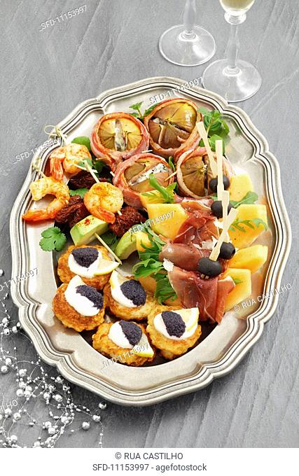 A platter of appetisers for Christmas, with melon, ham, potato fritters with caviar, figs with blue cheese, and prawn skewers